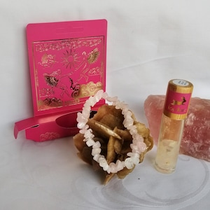 Puede incluir: Una pulsera de cuentas de virutas de cuarzo rosa descansa sobre una formación de cristal de rosa del desierto. También hay una caja rosa con gráficos de ángeles en pan de oro y un frasco roll-on con una etiqueta rosa. El fondo es blanco.