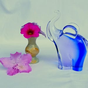 Könnte beinhalten: Eine klare und blaue Glas-Elefantenfigur mit gebogenem Rüssel und großen Ohren. Eine kleine Vase mit einer rosa Blume und eine lose rosa Blume sind ebenfalls im Bild. Der Elefant ist ein dekorativer Gegenstand.