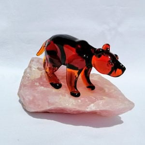 Puede incluir: Una pequeña figura de oso de vidrio translúcido con un degradado de colores ámbar oscuro y negro. El oso está de pie sobre un trozo rugoso e irregular de cristal de cuarzo rosa pálido, mostrando un acabado suave y pulido en el vidrio.