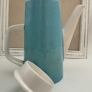 May include: A tall ceramic coffee pot with a glossy turquoise body featuring a fine crackle glaze finish. The pot has a white handle, a white spout, and a separate white lid with a small knob handle resting in front of the main vessel.