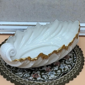 May include: A white ceramic bowl shaped like a large leaf with a curled stem at one end. The edges of the leaf-shaped bowl are detailed with a metallic gold trim that follows the scalloped contours of the rim, highlighting the textured veins.