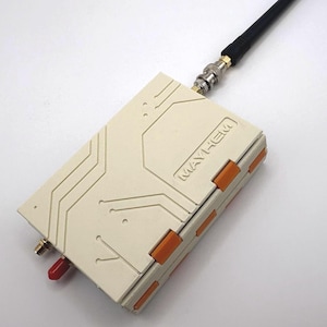 May include: A cream-colored electronic device with orange accents and the word "MAYHEM" printed on the side. It has a red antenna and a black cable with a silver connector attached. The device features circuit board-like detailing.