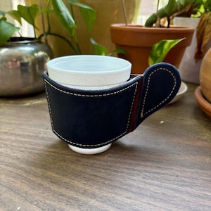 May include: A navy blue leather plant pot holder with a brown accent and white stitching. The holder has a handle and is designed to hold a white plant pot. The background includes other plants and pots.
