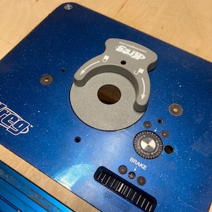 May include: A blue router table with a gray router lift and a brake dial. The router lift has the brand name "Kreg" printed on it. The table also has a slot for adjustments.