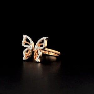 May include: A rose gold butterfly ring adorned with white gemstones. The butterfly wings feature small, sparkling stones, and the band is polished rose gold. The ring is displayed against a black backdrop.