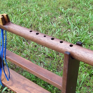 May include: A dark brown wooden Native American style flute with six finger holes and a carved block secured by a bright blue leather cord, resting horizontally on a matching dark brown wooden stand with two vertical supports and a base.