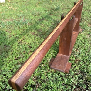 May include: A long, straight wooden staff featuring a multi-toned design with dark brown and lighter tan wood grain segments. The staff rests horizontally on a matching dark brown wooden stand that has two vertical supports and a flat base.