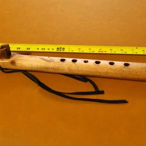 May include: A light brown wooden flute with six finger holes, a dark brown block attached to the top, and black leather ties. A yellow measuring tape placed above the flute indicates a total length of approximately 11.5 inches.
