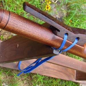 May include: A brown wooden Native American style flute featuring a dark brown block secured with a bright blue leather cord. The instrument rests on a matching brown wooden stand, showcasing a smooth polished finish and carved circular details.