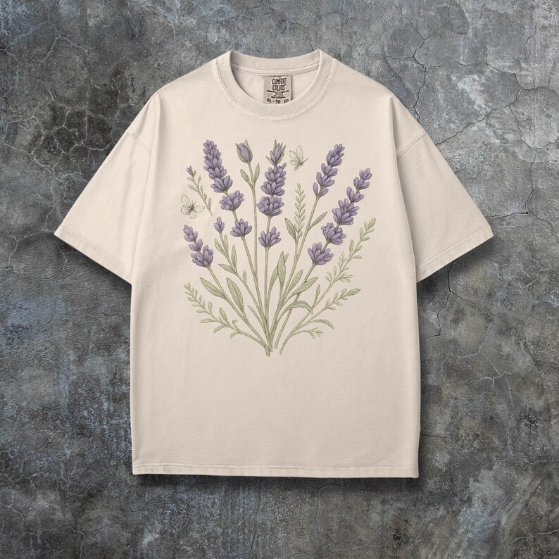 Puede incluir: Camiseta color crema con dise&ntilde;o de ramo de lavanda. La ilustraci&oacute;n incluye flores de lavanda moradas, hojas verdes y una peque&ntilde;a mariposa. La camiseta tiene cuello redondo cl&aacute;sico y mangas cortas, ideal para uso casual.