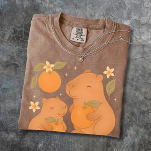 May include: A brown Comfort Colors t-shirt with a capybara family design, oranges, and floral details. The shirt has a crew neck and short sleeves. A silver necklace with a small pendant is draped across the shirt.