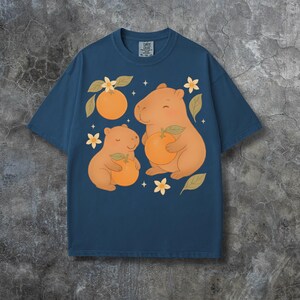May include: A navy blue t-shirt with a playful design of two capybaras, each holding an orange, with orange blossoms and leaves. The shirt has a crew neck and a relaxed fit. The tag reads "Comfort Colors".