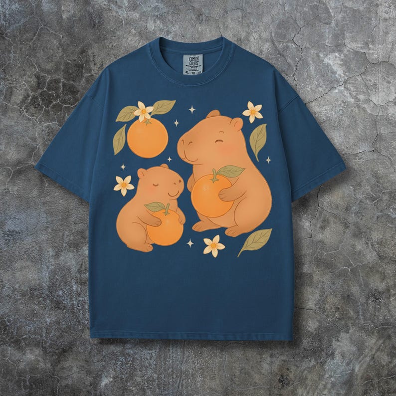 May include: A navy blue t-shirt with a playful design of two capybaras, each holding an orange, with orange blossoms and leaves. The shirt has a crew neck and a relaxed fit. The tag reads "Comfort Colors".