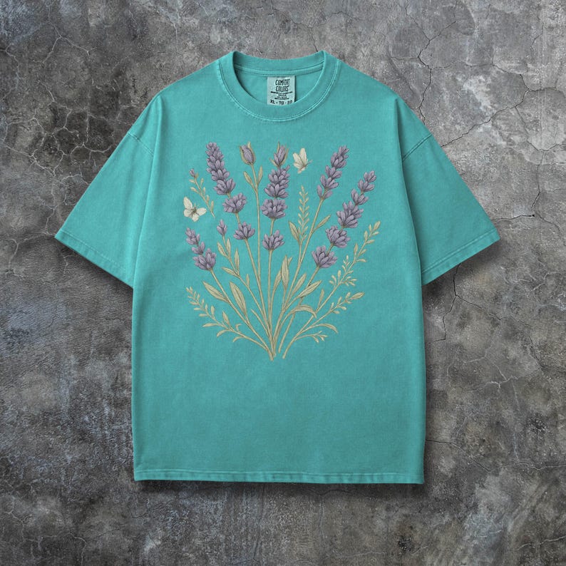 Puede incluir: Una camiseta turquesa con un dise&ntilde;o floral de lavanda y mariposas. La lavanda es morada, con tallos y hojas verdes. Tambi&eacute;n se incluyen dos mariposas en el dise&ntilde;o. La camiseta es de la marca Comfort Colors.