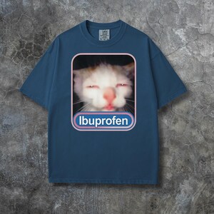 Puede incluir: Camiseta azul marino con un gr&aacute;fico de un gato con ojos rojos y la palabra "Ibuprofen" en un dise&ntilde;o blanco y azul. La camiseta tiene mangas cortas y cuello redondo.