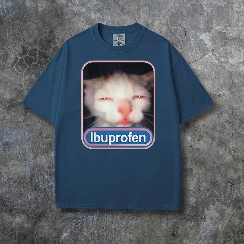 Puede incluir: Camiseta azul marino con un gr&aacute;fico de un gato con ojos rojos y la palabra "Ibuprofen" en un dise&ntilde;o blanco y azul. La camiseta tiene mangas cortas y cuello redondo.