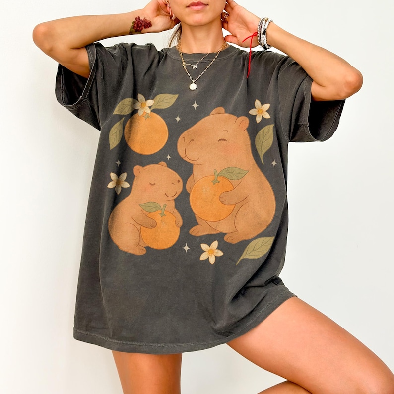 May include: Dark grey t-shirt with a playful design of two capybaras holding oranges, set amongst orange blossoms and leaves. The graphic is printed on a soft, oversized tee. The shirt has a vintage, washed appearance.