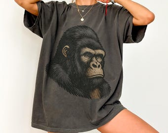 May include: Dark grey t-shirt featuring a detailed illustration of a gorilla's face. The gorilla is depicted in black and brown tones, with a serious expression. The shirt has short sleeves and a relaxed fit, suitable for casual wear.
