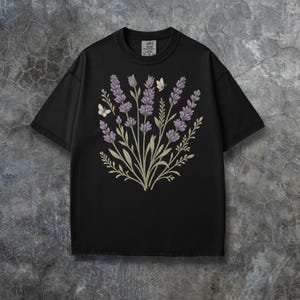 Puede incluir: Camiseta negra con un dise&ntilde;o floral de lavanda y dos mariposas. El dise&ntilde;o incluye flores de color lavanda, hojas verdes y tallos. La camiseta tiene cuello redondo y mangas cortas. La etiqueta de la camiseta dice "COMFY COLORS".