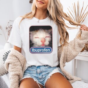 Puede incluir: Camiseta blanca con la cara de un gato con los ojos cerrados y la nariz rosa, enmarcada en un rect&aacute;ngulo rosa y azul. La palabra "Ibuprofeno" est&aacute; impresa debajo de la imagen del gato. La modelo lleva pantalones cortos vaqueros azules y un su&eacute;ter beige.