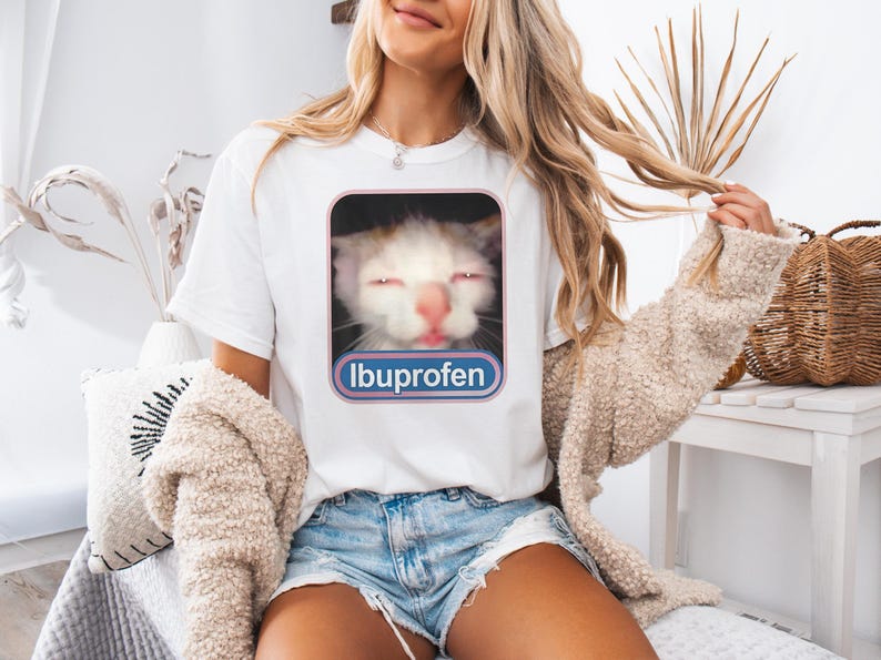 Puede incluir: Camiseta blanca con la cara de un gato con los ojos cerrados y la nariz rosa, enmarcada en un rect&aacute;ngulo rosa y azul. La palabra "Ibuprofeno" est&aacute; impresa debajo de la imagen del gato. La modelo lleva pantalones cortos vaqueros azules y un su&eacute;ter beige.