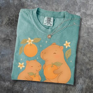 May include: A teal Comfort Colors t-shirt with a playful design of two capybaras holding oranges, surrounded by orange flowers and leaves. A silver necklace with a pendant is on the shirt. The tag reads "Comfort Colors".