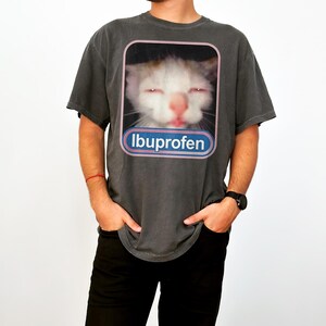 Puede incluir: Camiseta gris oscuro con un gato con los ojos cerrados y la palabra "Ibuprofeno" en un &oacute;valo azul y rosa. El gato tiene pelo blanco y naranja y una nariz rosa. La camiseta es de corte holgado.