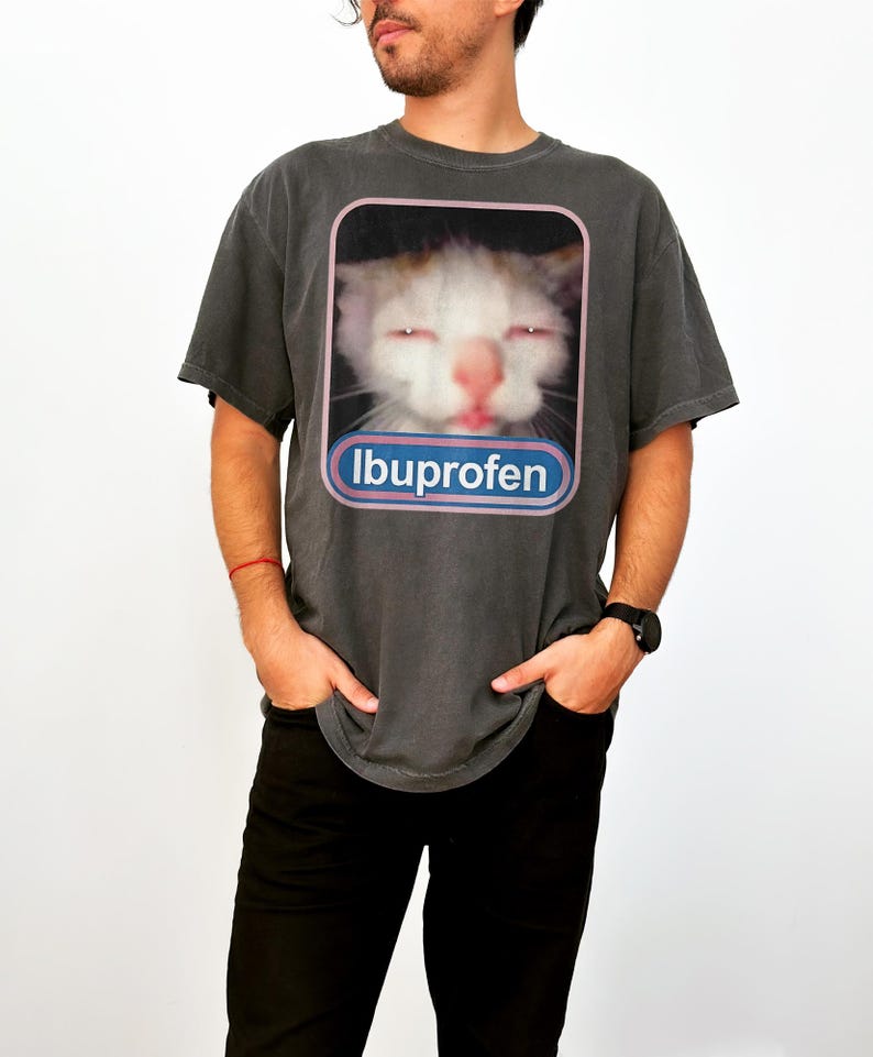 Puede incluir: Camiseta gris oscuro con un gato con los ojos cerrados y la palabra "Ibuprofeno" en un &oacute;valo azul y rosa. El gato tiene pelo blanco y naranja y una nariz rosa. La camiseta es de corte holgado.