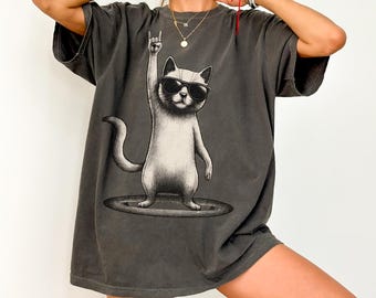 May include: Dark grey t-shirt featuring a cool cat wearing sunglasses and giving the rock on hand gesture. The cat is standing on a circular platform. The shirt has a vintage, washed look.