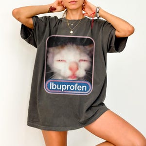 Puede incluir: Camiseta gris oscuro con un gr&aacute;fico de gato y la palabra "Ibuprofen" en un dise&ntilde;o azul y blanco. El gato tiene los ojos cerrados y la lengua fuera. La camiseta es de corte holgado.