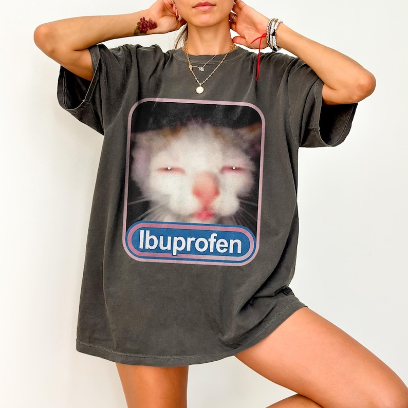 Puede incluir: Camiseta gris oscuro con un gr&aacute;fico de gato y la palabra "Ibuprofen" en un dise&ntilde;o azul y blanco. El gato tiene los ojos cerrados y la lengua fuera. La camiseta es de corte holgado.