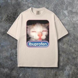 Puede incluir: Camiseta color crema con un gr&aacute;fico de la cara de un gato con ojos rojos y la palabra "Ibuprofeno" en un rect&aacute;ngulo redondeado azul y rosa. La camiseta tiene cuello redondo y mangas cortas.