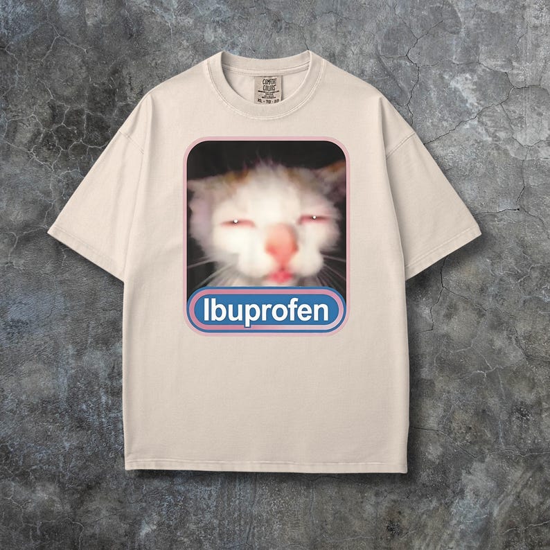 Puede incluir: Camiseta color crema con un gr&aacute;fico de la cara de un gato con ojos rojos y la palabra "Ibuprofeno" en un rect&aacute;ngulo redondeado azul y rosa. La camiseta tiene cuello redondo y mangas cortas.