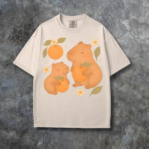 May include: A cream-coloured t-shirt with a playful design of two capybaras holding oranges, with orange blossoms and leaves. The shirt has a crew neck and short sleeves. The tag reads "COMFORT COLORS".