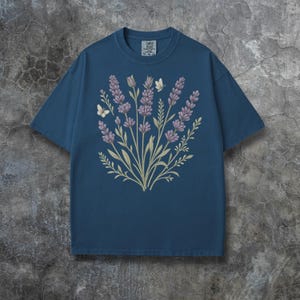 Puede incluir: Camiseta azul con un dise&ntilde;o floral de lavanda y mariposas. La lavanda es de color p&uacute;rpura suave, con tallos y hojas verdes. Dos mariposas blancas tambi&eacute;n est&aacute;n incluidas en el dise&ntilde;o. La camiseta tiene cuello redondo y mangas cortas.