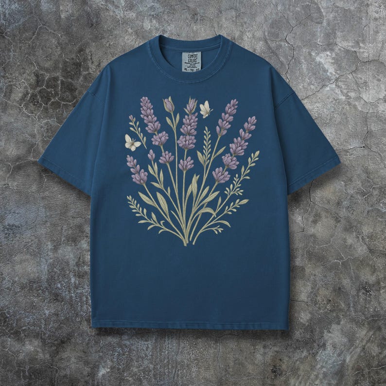 Puede incluir: Camiseta azul con un dise&ntilde;o floral de lavanda y mariposas. La lavanda es de color p&uacute;rpura suave, con tallos y hojas verdes. Dos mariposas blancas tambi&eacute;n est&aacute;n incluidas en el dise&ntilde;o. La camiseta tiene cuello redondo y mangas cortas.