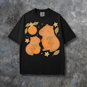 May include: Black t-shirt with a cartoon illustration of two capybaras holding oranges, with orange blossoms and leaves. The tag includes the text "COMFORT COLORS". A fun and playful design.