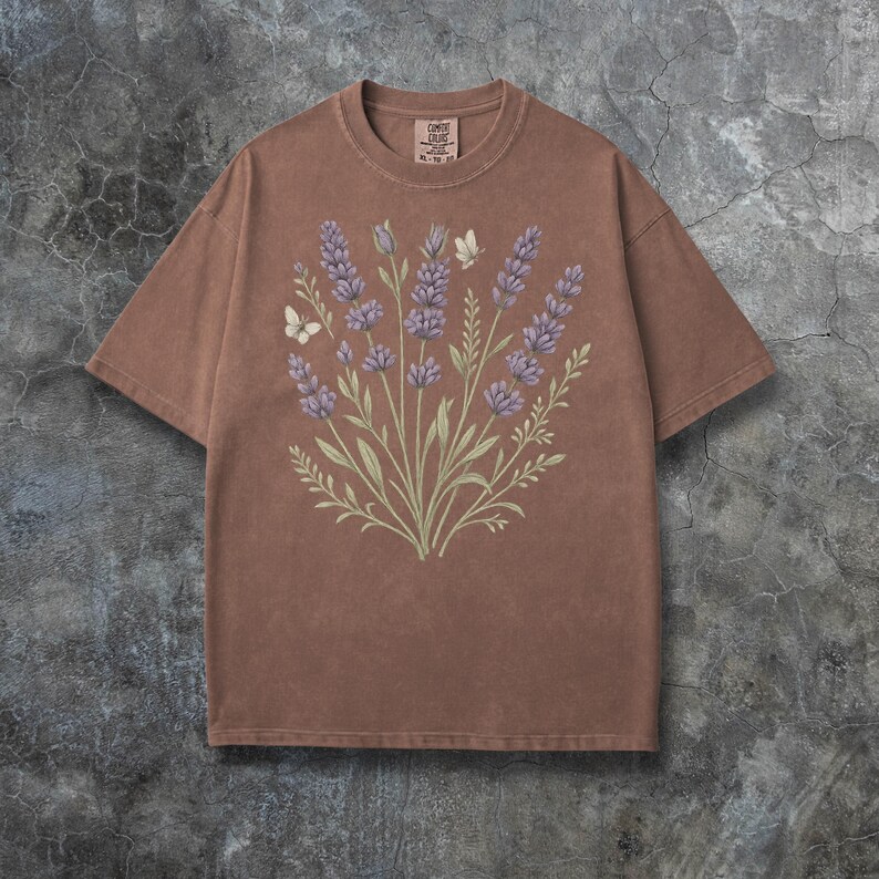 Puede incluir: Camiseta marr&oacute;n con un dise&ntilde;o de lavanda y mariposas. El gr&aacute;fico incluye ramitas de lavanda en tonos morados y verdes, con dos mariposas blancas. La camiseta tiene cuello redondo y mangas cortas. La etiqueta dice "Comfort Colors".