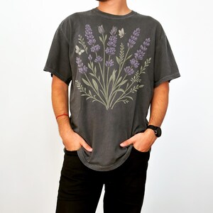 Puede incluir: Camiseta gris oscuro con un dise&ntilde;o de ramo de lavanda. El dise&ntilde;o incluye flores moradas, hojas verdes y dos mariposas. La camiseta tiene un corte holgado.