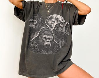 May include: Dark gray t-shirt featuring a graphic of three gorillas and a moon. The design is in black and white, with one gorilla roaring. The shirt has short sleeves and a relaxed fit.