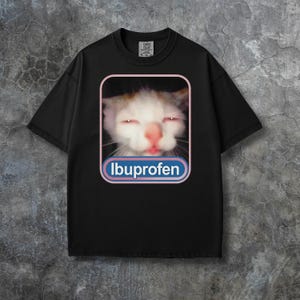 Puede incluir: Camiseta negra con un gato de ojos rojos y la palabra "Ibuprofeno" en un dise&ntilde;o azul y blanco. El gato tiene pelaje blanco y marr&oacute;n. La camiseta tiene cuello redondo y mangas cortas.