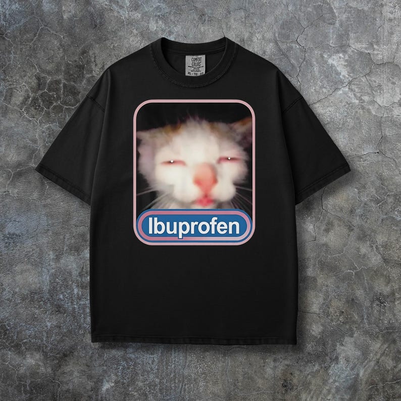 Puede incluir: Camiseta negra con un gato de ojos rojos y la palabra "Ibuprofeno" en un dise&ntilde;o azul y blanco. El gato tiene pelaje blanco y marr&oacute;n. La camiseta tiene cuello redondo y mangas cortas.