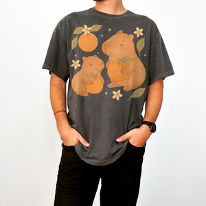 May include: Dark grey t-shirt with a playful design of two capybaras holding oranges, set amongst orange blossoms and leaves. The graphic is printed on a comfortable, oversized tee. A unique and fun graphic tee.