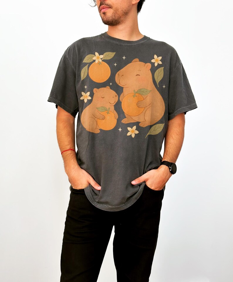 May include: Dark grey t-shirt with a playful design of two capybaras holding oranges, set amongst orange blossoms and leaves. The graphic is printed on a comfortable, oversized tee. A unique and fun graphic tee.