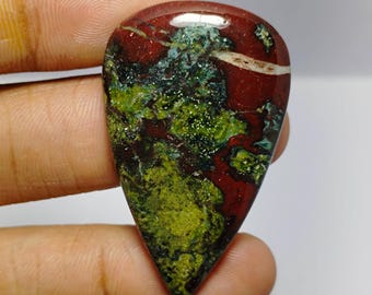 Könnte beinhalten: Ein polierter, tropfenförmiger Edelstein-Cabochon mit einem komplexen Muster aus tiefroten, leuchtend lindgrünen und dunkelwaldgrünen Mineraleinschlüssen sowie einer einzelnen dünnen weißen horizontalen Ader im oberen Bereich.