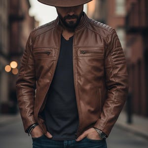 May include: A brown leather jacket with a quilted design on the shoulders and sleeves. The jacket has a zip-up front and two chest pockets. The person is wearing a brown hat, a black shirt, and blue jeans. A watch and bracelet are visible.