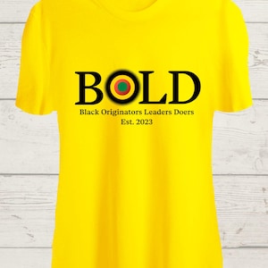 May include: A bright yellow t-shirt with the word "BOLD" in black, featuring a circular design with red, green, and yellow accents. Below the word "BOLD" is the text "Black Originators Leaders Doers Est. 2023".