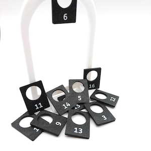May include: Black square tags with white numbers from 3 to 16, each with a hole at the top. A white arched display stand holds one tag. The tags are scattered on a white surface. The tags are likely for a game or organizational system.