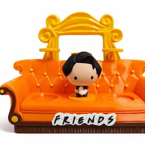 Könnte beinhalten: Ein orangefarbenes Miniatursofa mit einem gelben Bogen und einer Figur eines Mannes mit schwarzen Haaren. Auf dem Sofa steht das Wort "FRIENDS" in weißen Buchstaben. Das Sofa ist eine Nachbildung des Sofas aus der Fernsehserie.