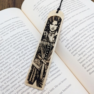 May include: Wooden bookmark featuring a detailed black and white illustration of a woman in a detailed outfit. The bookmark has a black string attached at the top and is placed on an open book.